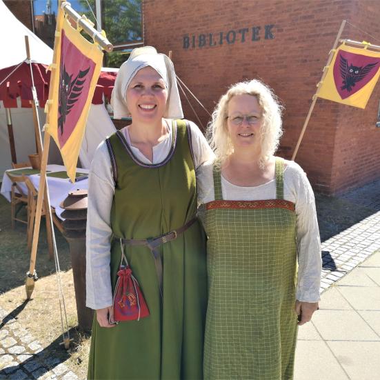 To biblioteksansatte står i middelalderdragter foran Nyborg Bibliotek og et gult flag med uglevåbenskjold.