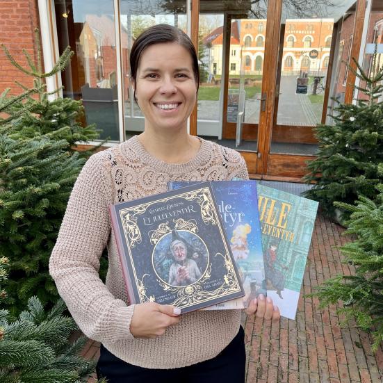 Marianne står omgivet af grantræer bag Nyborg Bibliotek, hvor hun holder de tre udgaver af "Et Juleeventyr", som hun anbefaler.