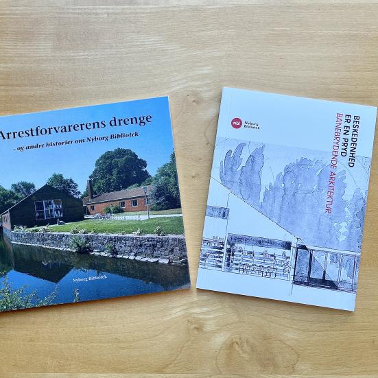 To bøger om Nyborg Bibliotek ligger på et lyst træbord.