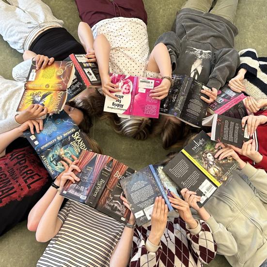 Readathon. Unge ligger i en cirkel med hovederne mod hinanden og ansigterne dækkede af åbne bøger.