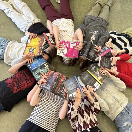 Readathon. Unge ligger i en cirkel med hovederne mod hinanden og ansigterne dækkede af åbne bøger.