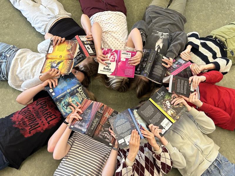 Readathon. Unge ligger i en cirkel med hovederne mod hinanden og ansigterne dækkede af åbne bøger.