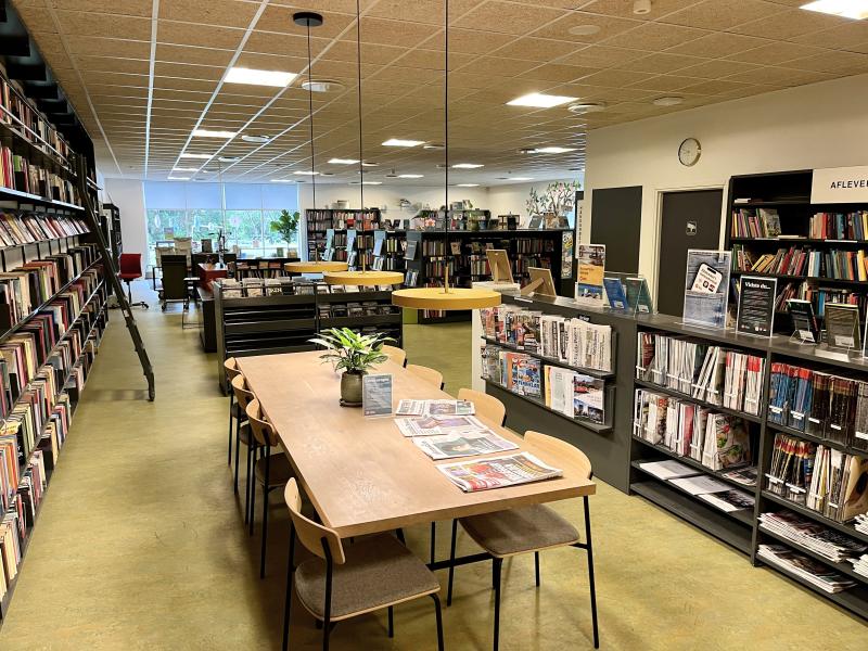 Billede af Ørbæk Bibliotek, der ligger i Ørbæk Midtpunkt. I forgrunden ses et bord med aviser. Til venstre en bogreol og i baggrunden flere bogreoler.