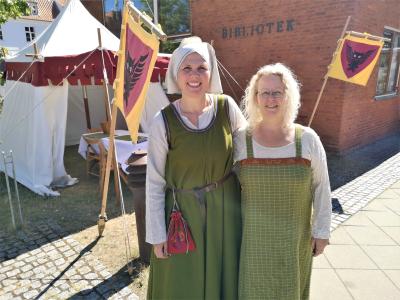 To biblioteksansatte står i middelalderdragter foran Nyborg Bibliotek og et gult flag med uglevåbenskjold.