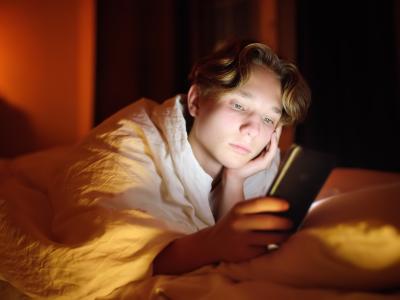 Unges digitale liv. Teenagedreng ligger i sin seng og kigger på sin mobil. Skærmen får hans ansigt til at lyse op. Hans ansigtsudtryk er meget neutralt.