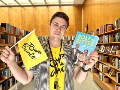Bibliotekar Mathias Lyngs står med Tour de France t-shirt og et Tour de France-flag og holder Rolf og Ritters bog.