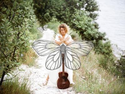 Billede af Stine Michel i naturen med en stor sommerfugl og en guitar. Fotograf: Signe Find Larsen.
