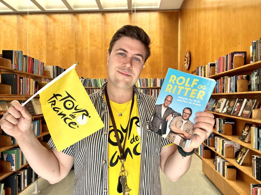 Bibliotekar Mathias Lyngs står med Tour de France t-shirt og et Tour de France-flag og holder Rolf og Ritters bog.