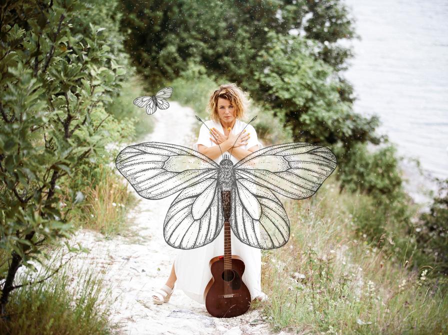 Billede af Stine Michel i naturen med en stor sommerfugl og en guitar. Fotograf: Signe Find Larsen.